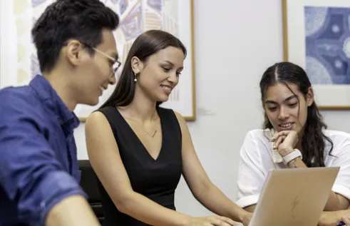 Three smiling people seated at a table around a laptop.